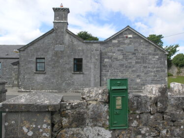 Limestone faced Scoil Naomh Bride, Tullach Ui Chadhain, built in 1862, with a post 2000 annex on right. A Victorian cast-iron post box (with VR initials) is set into the front wall. Royal Mail post boxes were painted bronze-green from 1859 until 1874 when the red colour became standard. Post boxes in Ireland were painted green after the Irish Free State was established in 1922.