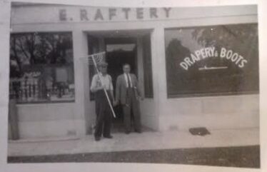 Two gentlemen in front of Rafter's shop, one holding what looks like a rake