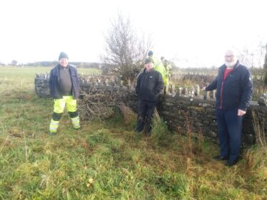 Three men standing by a stone wall in a green field