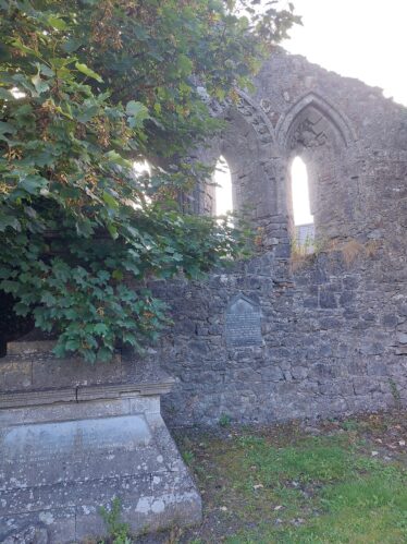 An old church wall is seen slightly obscured by a tree on the left side. The wall features 3 long narrow windows next to each other. They are all pointed on top. The base of a statue can also be seen on the left hand side but the rest of the figure is completely blocked by a the trees leaves. | James Spellman 2022
