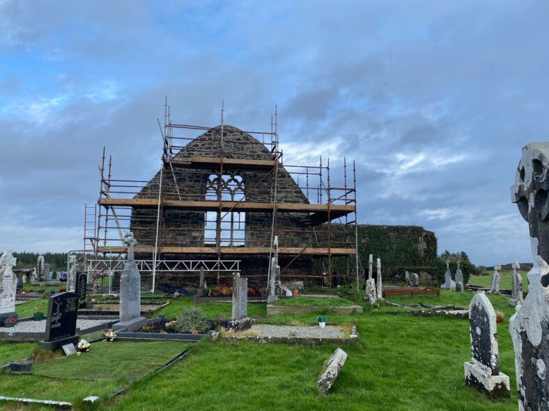 scaffold up partially blocking the tracery winfdow during works, blue sky anf green grass in foreground speckeld with gravemarkers | Fergal McNamara