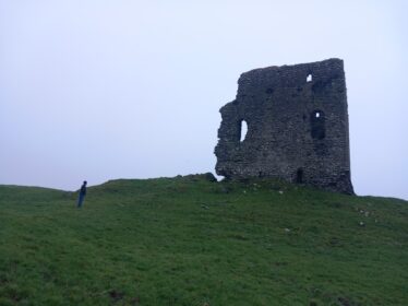 Moylough Casrle on a height, and a person looking up at it, green in foreground, grey sky behind