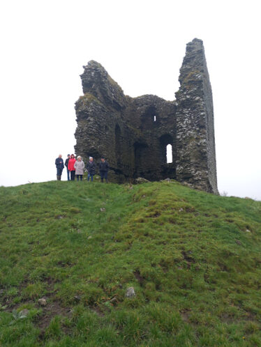 Moylough Castle on a height with a grey skey behind and a number of people standing beside the ruin. Green in the foreground | B. Doherty 2023