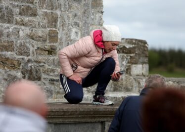 Girl wearing a coat and hat on her hunhers atop a chest tomb reading an inscription, with stone wall in background