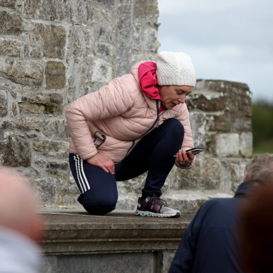 Girl wearing a coat and hat on her hunhers atop a chest tomb reading an inscription, with stone wall in background | Hany Marzouk