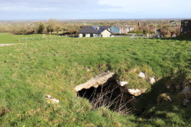 Rocks at entrance to souterrain within a green mound. Houses in the background | B. Doherty 2023