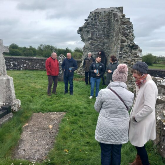 Two ladies in white coat look on, stone wall in background, graveslab on the ground within green grass | Marie Mannion