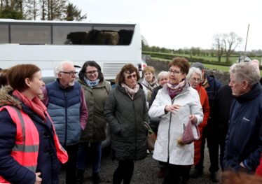 People n fron of a big white bus listens to lady talking