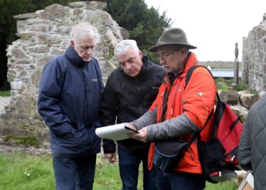 Man in hat shows two others a drawing at kilboght with church ruin in background