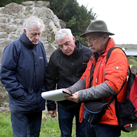 Man in hat shows two others a drawing at kilboght with church ruin in background