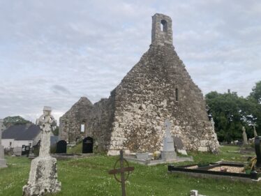 Gable end of church showing the bell tower on the apex located within a graveyard, pale blue sky with stringy white cloud in background, green grass in foreground dotted with memorials