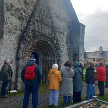 A group of people standing outside a Romanesque doorway as a man explains the history of it.