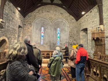 A group of people gathered inside Clonfert Cathedral looking at windows behind the altar. There are two window lights , stain glass in the background
