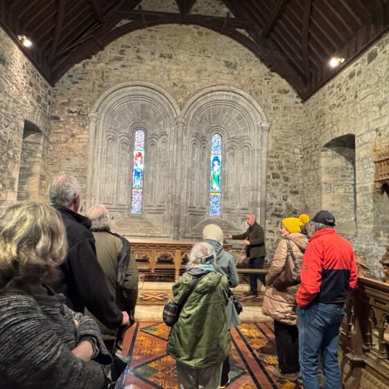 A group of people gathered inside Clonfert Cathedral looking at windows behind the altar. There are two window lights , stain glass in the background | Lucy Flynn 2026