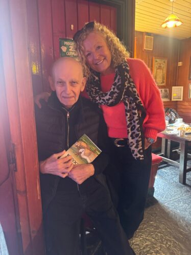An old man sitting while holding the book: Kilboght while a woman poses beside him for the picture. | Lucy Flynn 2026