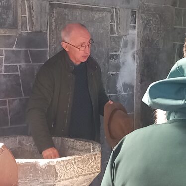 Man standing net to a bowl on a pedestal with his hand in it as he talks to a group of people.