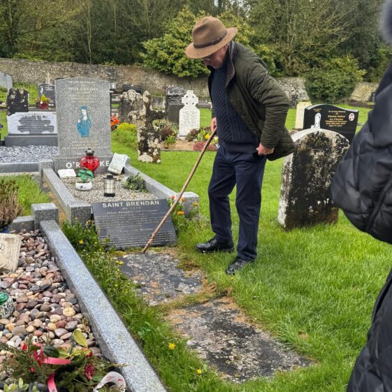 A man with a hat standing beside a grave, explaining the story of the deceased. | Lucy Flynn 2026