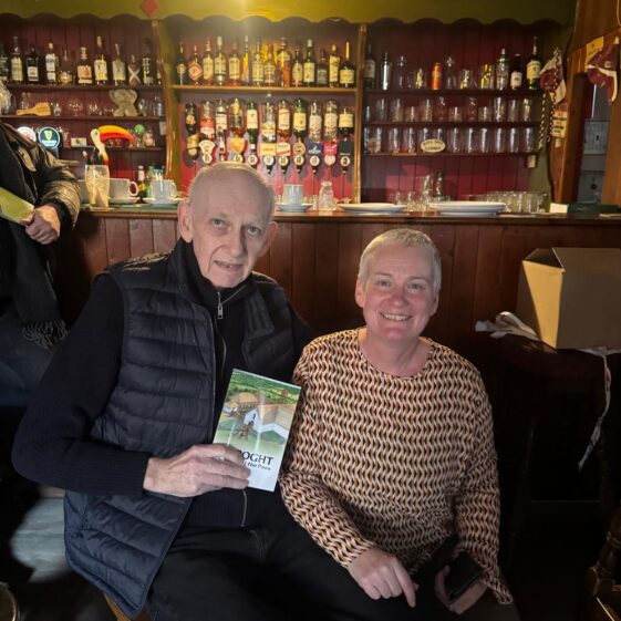 an old man sitting posing with a book while a lady squats beside him, both posing for the picture. | Lucy Flynn 2026
