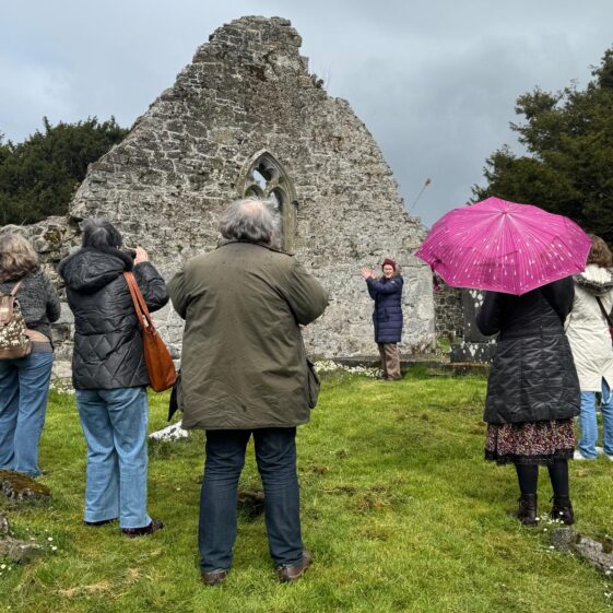 Group of people standing around the ruins of what is left of kilboght Church as a lady explains the history to the group. | Lucy Flynn 2026