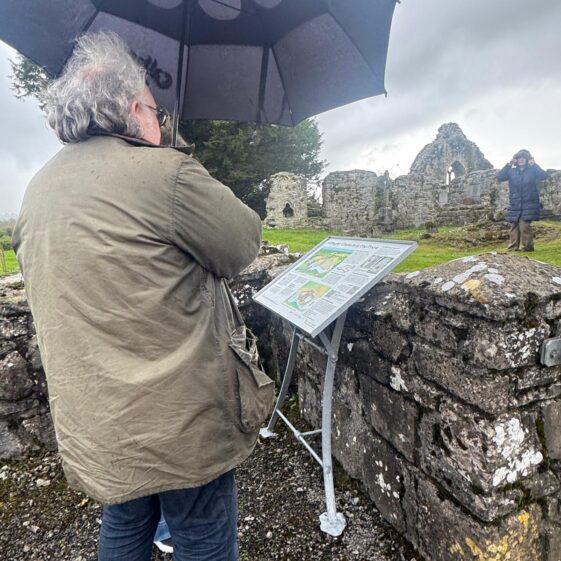 A man looking at a sign about the Kilboght ruins while holding an umbrella. | Lucy Flynn 2026