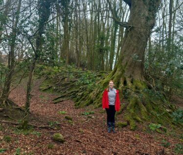A girl standing beside a tree that roots have grown around to adapt the wall of a enclousure. | B. Doherty 2026