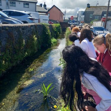 Children looking over a wall at water flowing along medieval moat of loughrea. High wall visisible on the left hand side, some greenery in the foreground | B. Doherty 2026