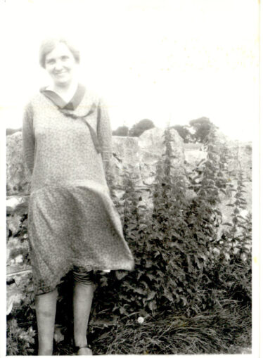 MAy Cregg smiling by a stone wall with flowers growing in background.