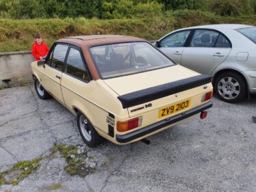 Old car parked in carpark with young boy admiring it.