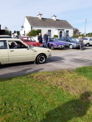 Green forwground, white, red and blue vintage cars parked in front of a cottage in Glentane, blue sky. People walking around.